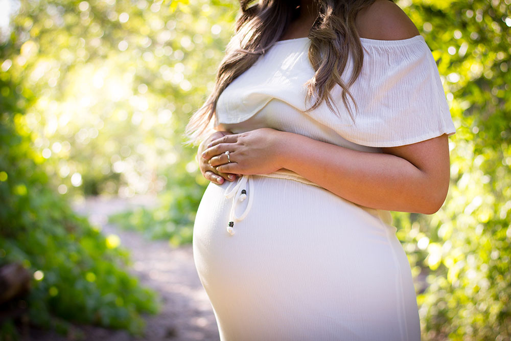 A pregnant white woman in a white dress, a ring on the finger. Some bush in the background