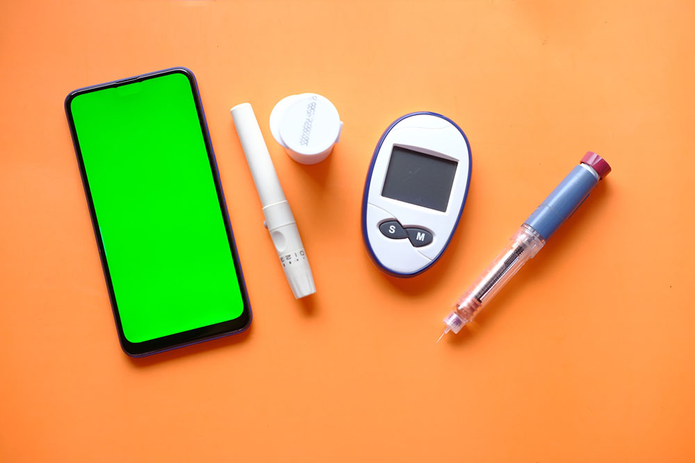 Photo or composition. A cell phone and a blood glucose meter kit (two cylindrical objects, a round jar and the monitor with two buttons). Solid orange background.