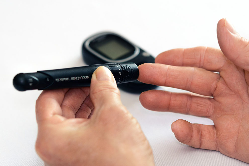 Photo: a hand holding a glucose meter and pointing to a finger of the other hand. In the background, a glucose monitor.