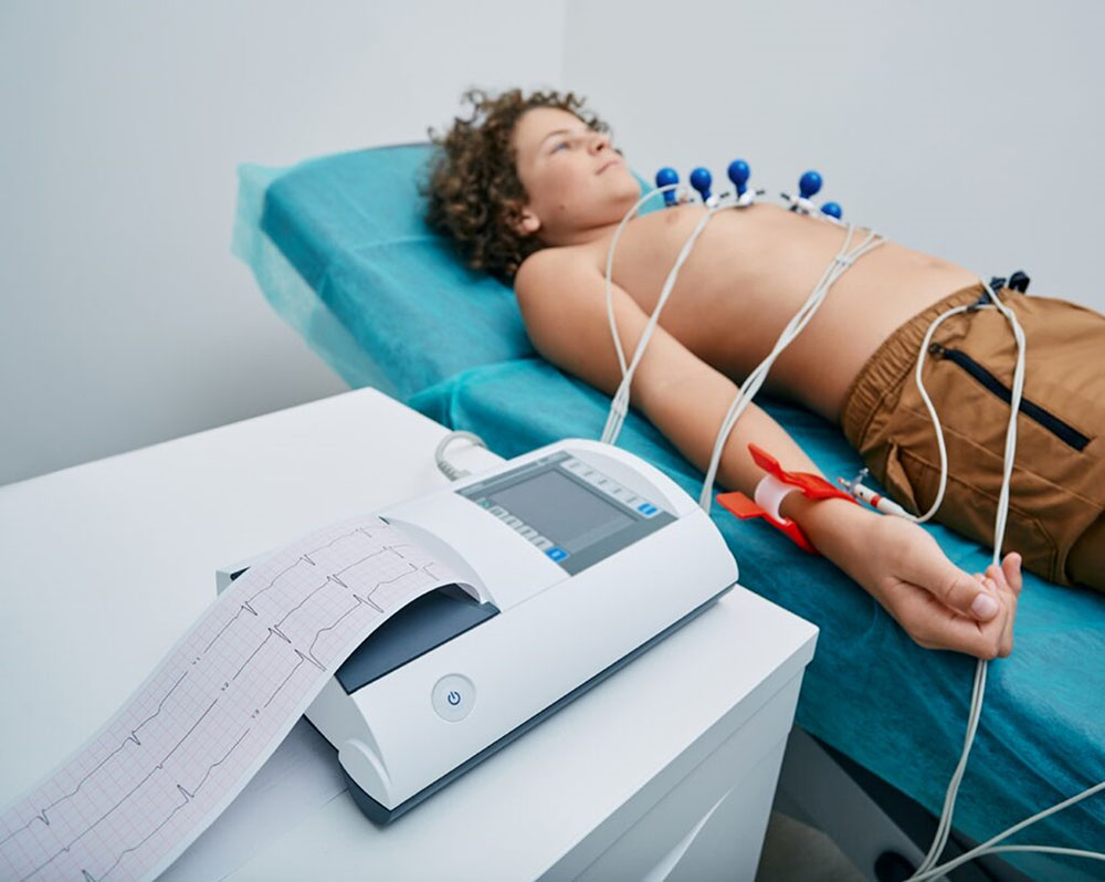Photo of a person lying on a hospital bed. He has no shirt and electrodes attached to his body. Next to the stretcher, an electrocardiograph producing a paper with the patient's heartbeat.