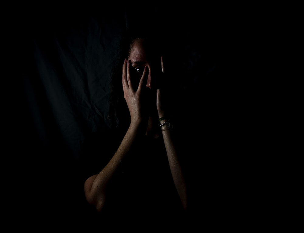 Foto com muita sombra, é possível ver apenas os reflexos na pele de uma mulher com as mãos no rosto. A imagem traz um sentimento de medo e solidão.