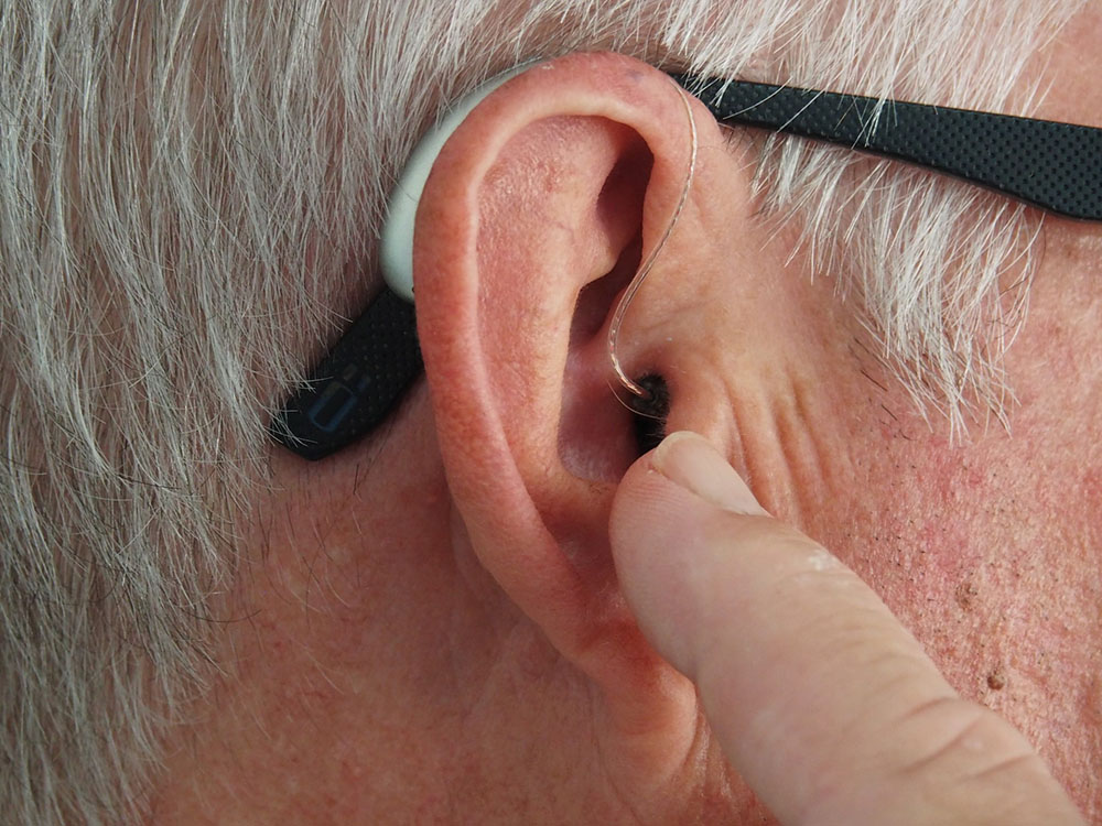 Foto: orelha de um homem idoso com aparelho de audição. Um dedo está apontando para o ouvido.