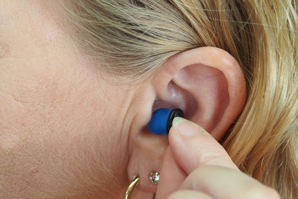 Foto: orelha de uma mulher loira com aparelho de audição. Ela está com os dedos em forma de pinça colocando ou tirando o dispositivo do ouvido.