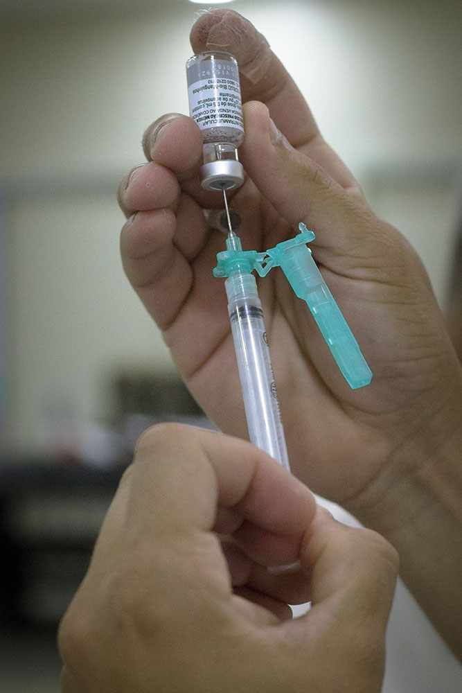 Foto: Duas mãos, uma segura um frasco de vacina e a outra uma seringa. A pessoa está transferindo o conteúdo do frasco para a seringa.