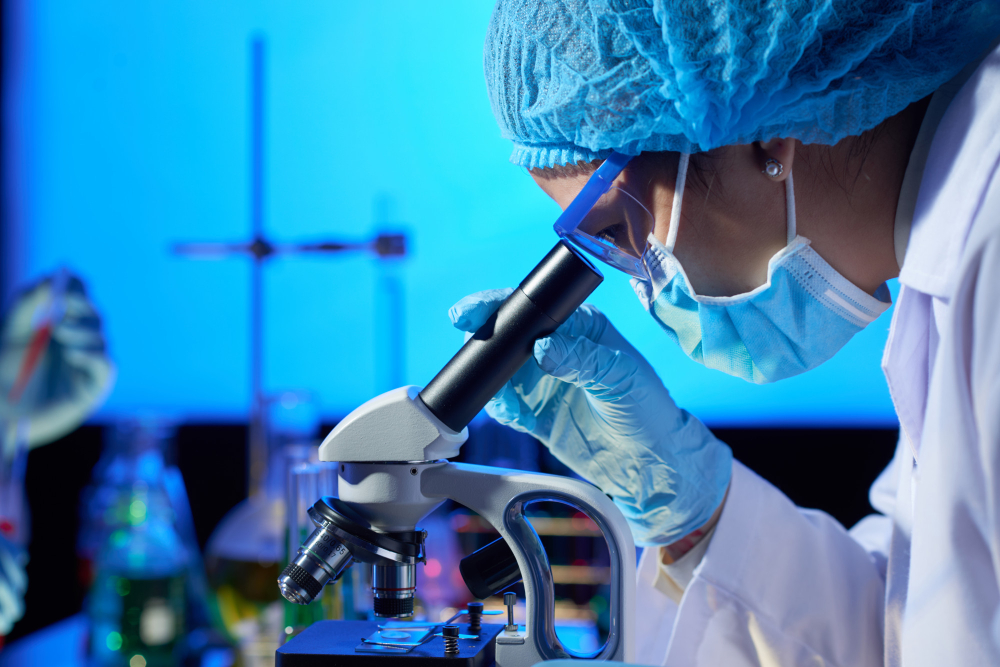 Foto de uma mulher olhando no microscópio. Ela usa óculos, jaleco, touca, máscara e luvas. No fundo, recipientes típicos de um laboratório.