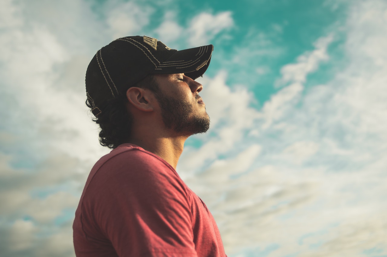 Foto de um homem de olhos fechados, expressão calma e cabeça erguida. No fundo, céu azul com nuvens.