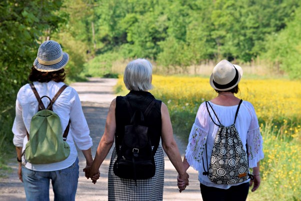 Foto: três mulheres de costas, andando em uma trilha de mãos dadas. No fundo, um espaço verde com árvores e grama.