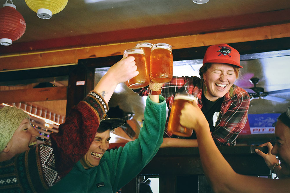 Foto: grupo de quatro mulheres comemorando, brindando canecas de vidro cheias de cerveja. O lugar parece ser um bar.