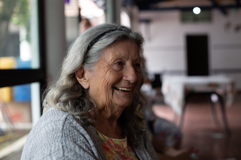 Foto de uma mulher idosa, com cabelos grisalhos na altura dos ombros, sorrindo. Fundo desfocado.