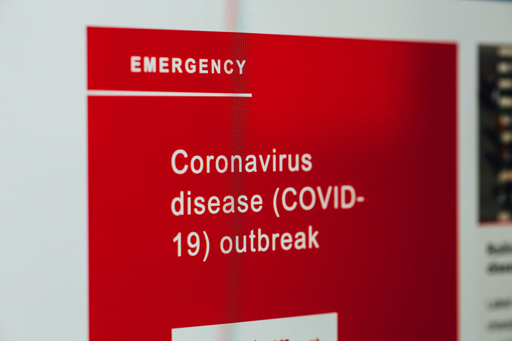 Fotografia de uma placa de emergência nas cores vermelho e branco, onde está escrito "Coronavírus disease (COVID-19) outbreak". Em português:"Surto da doença do coronavírus (COVID-19)".