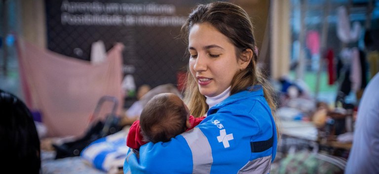 Fotografia de uma pessoa vestindo um uniforme azul do SUS segurando um bebê nos braços. Fotografia de uma pessoa vestindo um uniforme azul do SUS segurando um bebê nos braços.