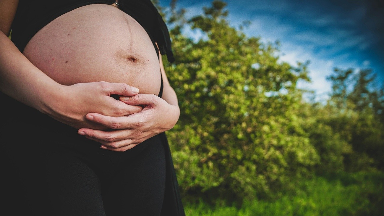 Uma fotografia de perto retrata uma pessoa grávida, com as mãos segurando a barriga, em um ambiente ao ar livre. Uma fotografia de perto retrata uma pessoa grávida, com as mãos segurando a barriga, em um ambiente ao ar livre.