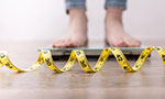 A yellow tape measure spirals over a wooden floor. Behind it the feet of a white-skinned person on top of a bathroom scale.