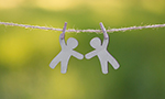 Photograph of two paper cutouts shaped like human figures, clipped to a string with clothespins and hanging against a blurred green background.
