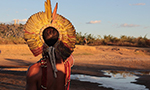 Photograph of an indigenous man seen from behind, gazing at the horizon. He is wearing a feather headdress and various adornments on his body. He stands beside a small lake in an open field with diverse vegetation. The sky above is clear and blue.