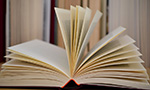 Photo of an open book on a table, in the background there's a bookcase full of books