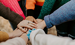 Fotografía de un grupo de personas, cada una con suéteres de diferentes colores, coloca sus manos juntas en el centro en señal de unidad.