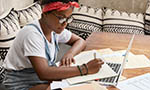 Sideways shot of black busy female journalist studies papers for writing article, uses online issue on laptop computer, sits at wooden desk, writes something down, wears denimm sarafan and spectacles