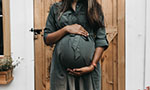 Foto de uma mulher negra grávida usando vestido. No fundo uma porta de madeira.