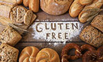 Foto: diferentes tipos de pães dispostos em volta de um punhado de farinha branca espalhado sobre uma mesa de madeira escura. Sobre os pães, dois ramos de trigo. Na farinha, as palavras "gluten free" foi desenhada.