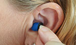 Foto: orelha de uma mulher loira com aparelho de audição. Ela está com os dedos em forma de pinça colocando ou tirando o dispositivo do ouvido.