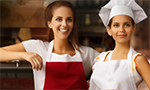 Imagem produzida com inteligência artificial: duas mulheres com avental, uma delas usa chapéu de chef de cozinha. A mulher da esquerda está sorrindo, a da direita tem uma expressão neutra. Na frente, uma bancada e duas pizzas.