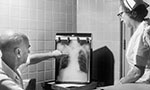 Foto em preto e branco. Um homem e uma mulher em uma sala. O homem aponta para um quadro com uma radiografia de tórax.