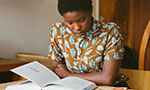 Foto de uma mulher com expressão neutra lendo um livro. Parece que ela está estudando, pois tem marcadores coloridos nas páginas do livro e um caderno do lado. Ela está com os braços apoiados em uma mesa de madeira.