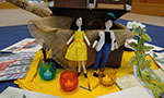 Foto. Duas bonecas de pano na frente de um baú. Na frente das bonecas, três velas coloridas sobre um pano amarelo. Ao redor do baú, das bonecas e das velas, vários desenhos.