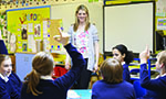 Foto de uma sala de aula. A professora está em pé sorrindo para os alunos. Dois deles estão com uma das mãos levantadas.