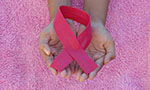 Foto. Laço cor de rosa da campanha de conscientização do câncer de mama. Uma pessoa segura o laço com as duas mãos juntas e viradas para cima. No fundo, um tecido de pelúcia rosa.