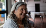 Foto de uma mulher idosa, com cabelos grisalhos na altura dos ombros, sorrindo. Fundo desfocado.