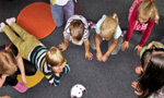 Foto de crianças brincando no chão, em um carpete cinza, acompanhados de uma adulta.