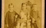Três crianças: uma menina está no centro, sentada e segurando um brinquedo, enquanto dois meninos estão em pé, um em cada lado dela.