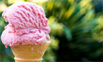 Mão segurando uma casquinha de sorvete. A massa do sorvete tem a cor rosa. O fundo da imagem é verde e está desfocado.