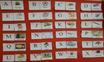 Painel vermelho com um alfabeto, onde, ao lado de cada letra, há uma representação visual de um objeto cujo nome começa com a mesma letra do alfabeto.