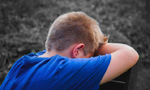 Um menino loiro, vestindo uma camiseta azul, está sentado de costas, com o rosto escondido entre os braços, possivelmente indicando que está triste.
