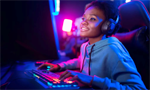 Mulher negra, de perfil, com fone de ouvido e um moletom cinza, em uma sala gamer. Ela utiliza um teclado com luzes de neon, concentrada diante de um monitor não visível na foto.