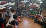Professora e alunos sentados no chão de uma sala de aula, em círculo. A docente está sorrindo, com um livro em mãos.