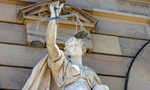 Escultura de Têmis (deusa grega da justiça) exibindo uma expressão destemida enquanto segurando uma balança, símbolo da justiça, em frente a um edifício. Ela está ladeada por duas crianças, um menino e uma menina, como se as estivesse protegendo.