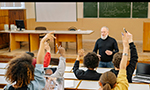 Professor de meia-idade ensinando em uma sala de aula ampla, onde alunos estão sentados em suas carteiras, com notebooks abertos diante deles, e vários levantam as mãos para fazer perguntas
