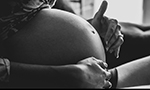 Fotografia em preto e branco de uma mulher grávida sentada, com a barriga à mostra e as mãos repousando sobre ela.