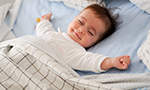 Bebê deitado em uma cama, com lençol azul-bebê, fronhas e edredom branco com quadriculado preto. Ele está de olhos fechados, sorrindo e com os braços abertos, transmitindo uma sensação de conforto e serenidade.