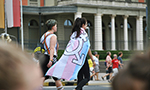 Duas pessoas vistas de perfil caminham em uma manifestação de rua, uma delas carregando uma grande bandeira trans. Outras pessoas, desfocadas, podem ser vistas ao fundo.
