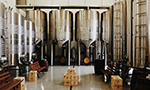 Fotografia do interior espaçoso de uma cervejaria ou vinícola com grandes tanques de fermentação de aço inoxidável reluzentes, caixas de madeira e bancos.