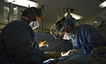 Fotografia de dois cirurgiões na sala de cirurgia realizando um procedimento. Eles estão usando máscaras cirúrgicas, aventais e focos de luz. Equipamentos médicos e bolsas de soro são visíveis ao fundo.