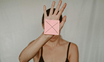 Fotografia de uma mulher com uma regata preta, levantando a mão com um post-it rosa colado, que tem um "X" preto desenhado, posicionado à frente do rosto.