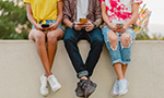 Fotografia de três pessoas sentadas em um muro, segurando seus smartphones. A imagem está cortada e não permite que elas sejam vistas do ombro para cima. Elas estão vestindo roupas casuais: uma camisa amarela, uma camisa xadrez e uma camisa vermelha tie-dye com jeans e tênis.