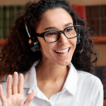 Fotografia de uma mulher à frente de um notebook usando um headset e acenando para a câmera do computador