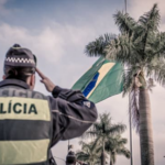 Fotografia de uma pessoa com uma farda da polícia de costas para a câmera batendo continência para a bandeira do Brasil do um coqueiro ao fundo